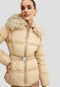 Beige pufferjakke med faux fur-krave, bælte i taljen, kontrasterende syninger og en sølvfarvet lynlås med en cirkulær guldfarvet detalje.