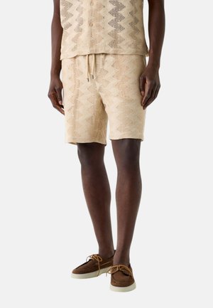 Uomo che indossa pantaloncini in maglia beige con motivo a fantasia abbinati a una camicia con bottoni e scarpe casual marroni in pelle con lacci, in piedi contro uno sfondo bianco.