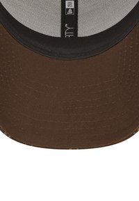 Ruskea lippalakki mustalla lipalla, jossa on verkkomateriaali sisäpuolella ja ompeluyksityiskohtia. Alapuolella on hienovarainen tekstuurikuvio.