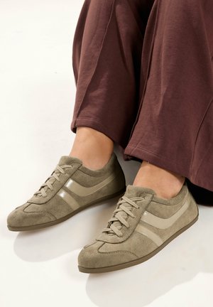 Stopy w lekkich brązowych zamszowych sneakersach z beżowymi paskami i sznurówkami, w połączeniu z luźnymi brązowymi spodniami na neutralnym tle.