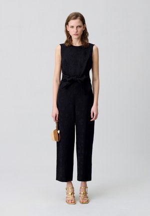Combinaison noire sans manches avec une taille à nouer, arborant un motif floral texturé. Associée à des sandales dorées à lanières et un petit sac marron.