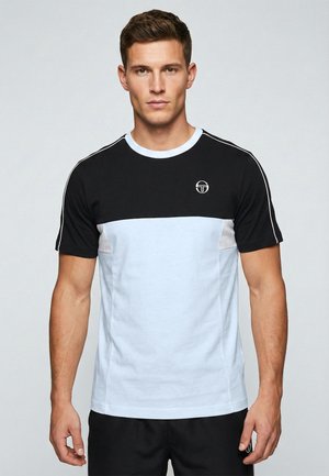 Homme aux cheveux courts et bruns portant un t-shirt noir et blanc à manches courtes avec un petit logo circulaire sur la poitrine.