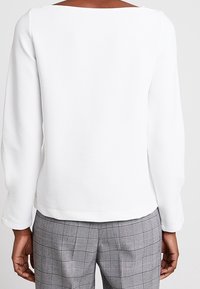 Bílý dlouhý rukáv s čtvercovým výstřihem, hladký povrch a jednoduchý design, spárovaný se šedými kalhotami s kostkovaným vzorem.