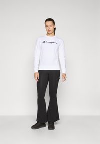 Vit sweatshirt med en svart "Champion"-logotyp, kombinerad med svarta utsvängda byxor och svarta sneakers. Enkel design med en ren estetik.