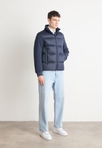 Ναυτική πούφφερ ζακέτα με φερμουάρ μπροστά, πλαϊνές τσέπες και ριχτές μανσέτες, συνδυασμένη με ανοιχτό μπλε τζιν και λευκά αθλητικά παπούτσια. Μινιμαλιστικός σχεδιασμός.