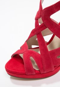 Sandale rouge en daim à talon haut avec des brides croisées et bout ouvert, montrant un matériau doux et texturé et une doublure intérieure beige.