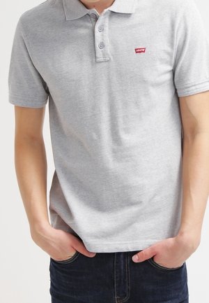 Person trägt ein hellgraues Levi's-Poloshirt mit zwei Knöpfen, Hände in den Taschen und dunkelblaue Jeans.