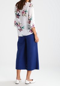 Bílá blůza s barevným potiskem motýlů, tříčtvrteční rukávy. Modré culottes a bílé nazouvací boty dotvářejí outfit.