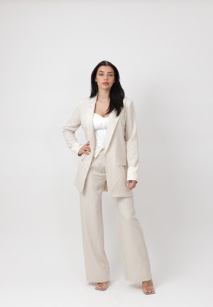 Kobieta o długich, ciemnych włosach stoi pewnie, ubrana w beżowe spodnie szerokie nogawki, pasującą marynarkę, biały top i sandały na obcasie na gołym tle.