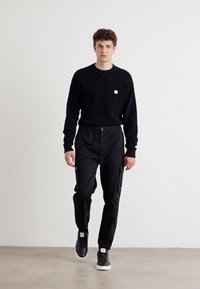 Zwart getextureerd sweatshirt met een klein logo, gecombineerd met zwarte cargobroeken met zijzakken, en zwarte sneakers met witte accenten.