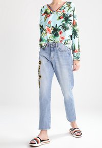 Květinový top s dlouhým rukávem v světle modré barvě, s tropickými potisky; světle džínové kalhoty s vyšitými akcenty; černobílé pruhované šlapky.