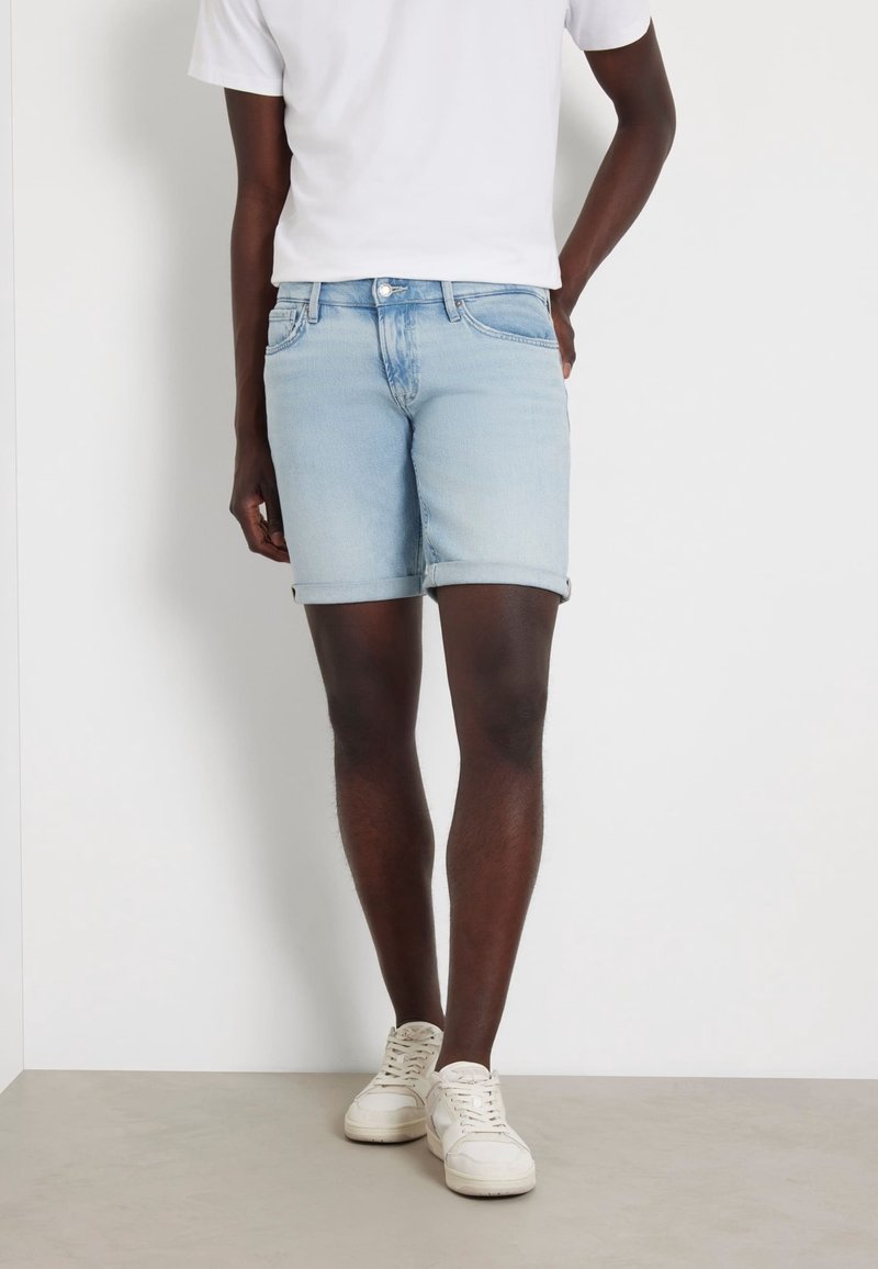 Personne portant un short en jean bleu clair, un t-shirt blanc et des baskets blanches, debout devant un mur et un sol blancs unis.