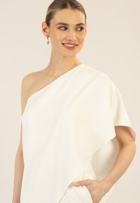 Haut blanc asymétrique avec une seule manche courte, tissu lisse et coupe décontractée. Comprend une poche cachée sur le côté.