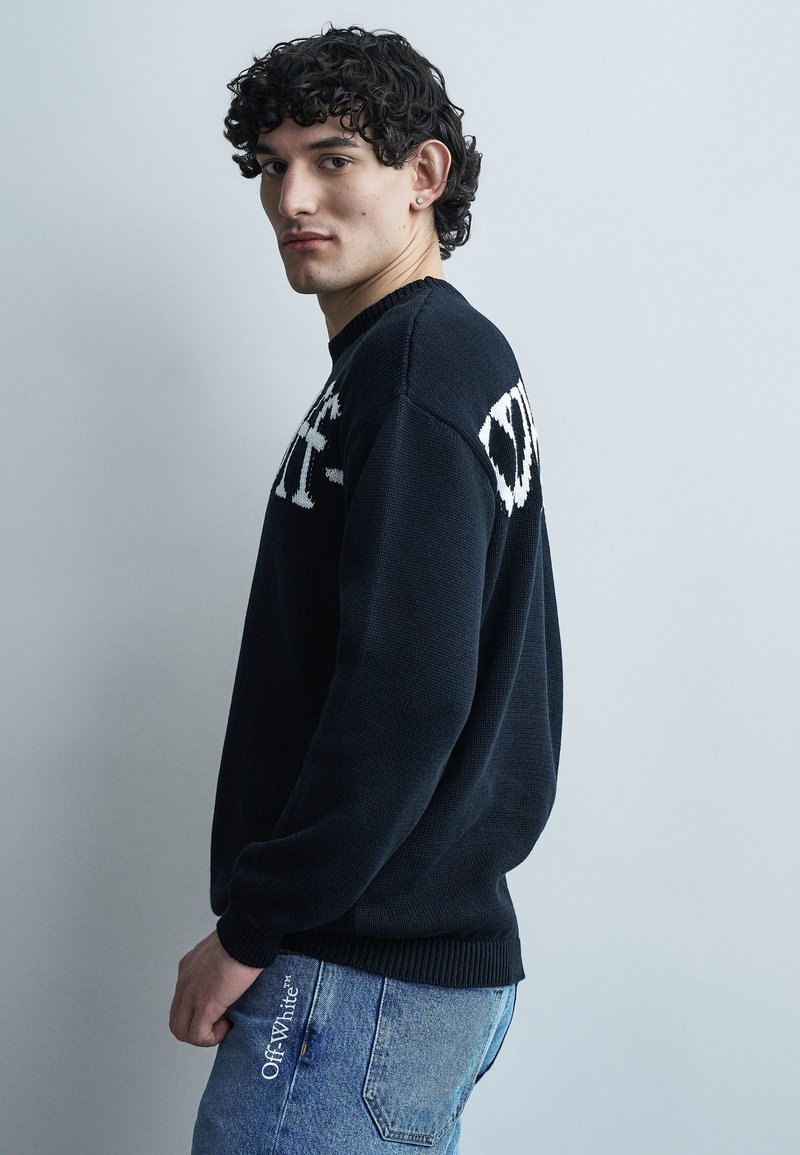 Νεαρός με σγουρά μαλλιά, που φοράει μαύρο πουλόβερ με λευκό κείμενο και ανοιχτό μπλε τζιν με λευκό λογότυπο "Off-White" στην τσέπη.