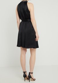 Femme portant une robe noire sans manches avec un col en perles, des escarpins noirs à bride de cheville, se tenant devant un fond clair et uni.