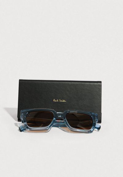 Kék átlátszó, téglalap alakú napszemüveg sötét lencsékkel, egy fekete bőr tokban, melyen aranyozott "Paul Smith" felirat található.