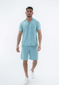 Chemise turquoise à manches courtes avec des boutons en bois et un short assorti, tous deux en tissu texturé. Porté avec des baskets blanches.