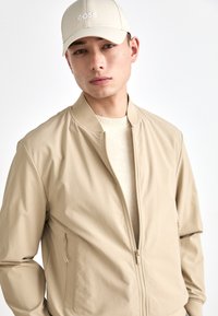 Beige bomberjakke med glidelås foran, ribbestrikket krage og sidelommer. Kombinert med en beige caps med hvit "BOSS" brodering.