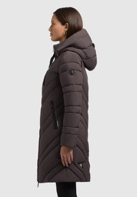 Donna che indossa un lungo cappotto invernale imbottito scuro con cappuccio, tasche con zip e toppe con logo, rivolta a sinistra in profilo su uno sfondo uniforme.