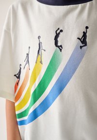 Fehér póló kerek nyakkivágással, színes átmenetes csíkokkal (piros, narancs, sárga, zöld, kék) és fekete kosárlabdázó sziluettekkel.