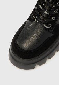 Bota de salto baixo preta com parte superior em couro texturizado, detalhes em camurça, biqueira arredondada e sola grossa de borracha. Atacadores com padrão ziguezague cinza.