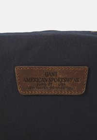 Etichetta in pelle marrone impressa con "GANT AMERICAN SPORTSWEAR 06516 CT – USA NEW HAVEN CONNECTICUT" su tessuto blu scuro.