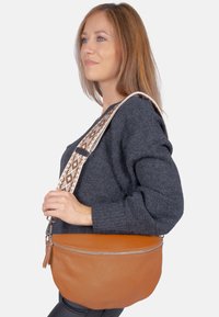 Sac à bandoulière en cuir marron avec une forme courbée, présentant une surface texturée et une sangle à motifs dans des tons beiges et marron.