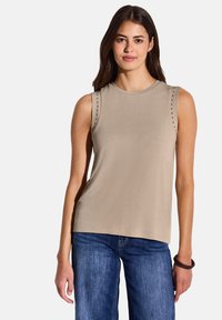 Top beige senza maniche con scollatura rotonda; presenta borchie decorative lungo le cuciture delle spalle. Abbinato a jeans blu, in tessuto semplice.