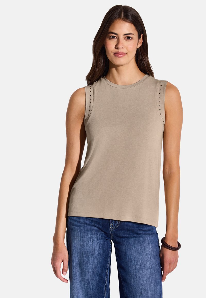 Top beige senza maniche con scollatura rotonda; presenta borchie decorative lungo le cuciture delle spalle. Abbinato a jeans blu, in tessuto semplice.