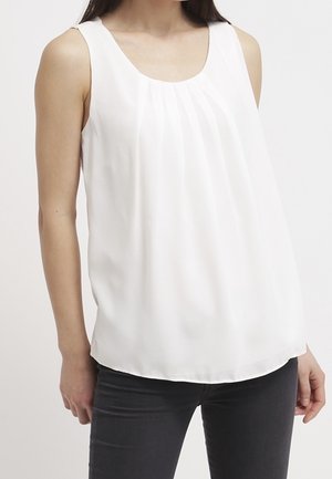 Femme portant un chemisier blanc plissé sans manches associé à un pantalon ajusté sombre, debout devant un fond clair uni.