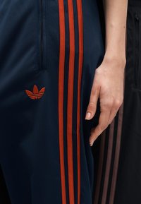 Χέρι που ακουμπάει δίπλα σε μπλε σκούρο παντελόνι Adidas με τρεις πορτοκαλί ρίγες και κεντημένο λογότυπο τριφυλλιού στο μηρό.