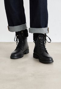 Black leather ankle boots with lace-up fronts and fur lining, worn with dark rolled-up jeans on a light floor.