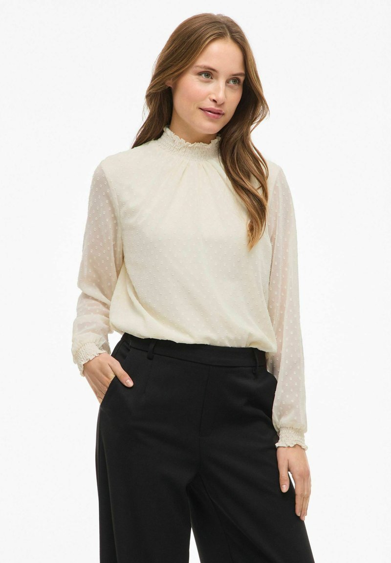 Cremebluse med en høj volangkrave, gennemsigtige lange ærmer og tekstureret polka dot mønster. Kombination med skræddersyede sorte bukser.