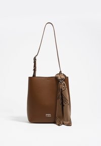 Braune Leder-Tote mit einem einzelnen Henkel, der mit einem dekorativen beigen Schal geschmückt ist, der am Henkel gebunden ist, sowie mit dezentem Branding am Boden.