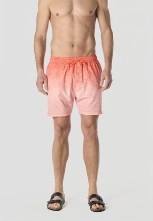 Hombre con bañador corto degradado coral con un sutil patrón de hojas, sandalias negras, de pie contra un fondo claro y sencillo.