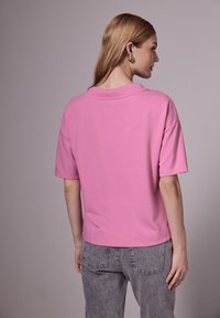 Růžová mikina s krátkým rukávem, volného střihu, kulatým výstřihem a měkkou texturou. Nosená s světle šedými džíny.