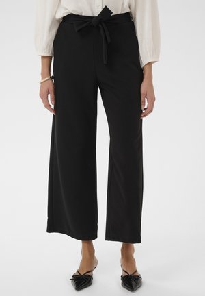 Žena nosi čierne široké nohavice s vysokým pásom a opaskom, v kombinácii s čiernymi špicatými balerínami a bielou blúzkou s dlhým rukávom.