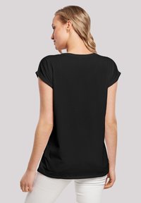 Schwarzes T-Shirt mit kurzen, umgeschlagenen Ärmeln, lässiger Schnitt und glatter Textur, getragen mit weißen Hosen, von hinten gesehen.