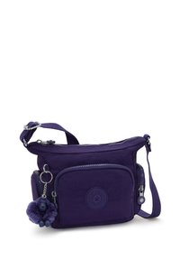 Malá fialová crossbody taška s přední kapsou na zip, nastavitelným popruhem, bočními přihrádkami a plyšovým klíčenkou opice připevněnou na zipu.