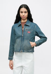 Νεαρή γυναίκα με μακριά σκούρα μαλλιά φοράει ένα μπλε μπουφάν από τζιν πάνω από ένα καφέ πουκάμισο και λευκό παντελόνι, στέκεται μπροστά σε απλό λευκό φόντο.