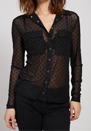 Woman wearing a sheer black long-sleeve blouse with polka dots and front pockets over a black top, paired with black pants.