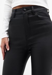 Pantalon noir en similicuir taille haute avec une texture lisse, doté de poches avant et de détails de coutures visibles.