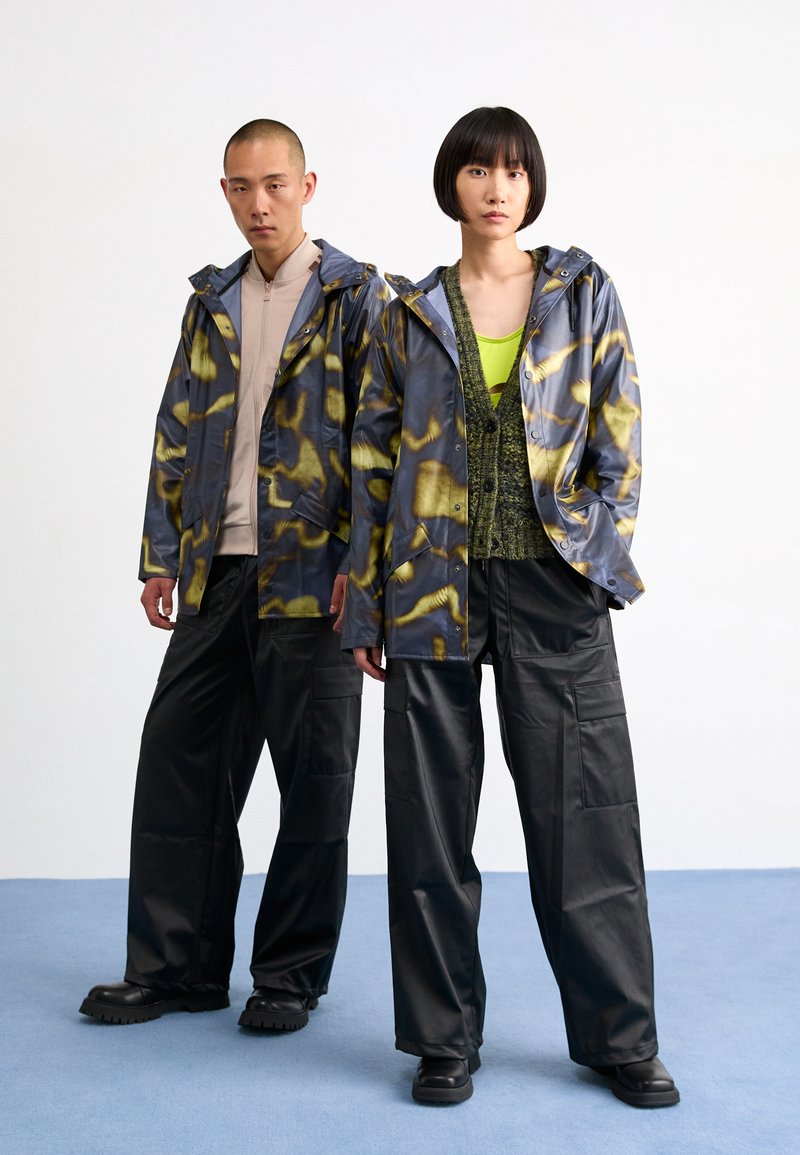 Deux individus portent des vestes imperméables à capuche à motifs bleus et dorés, associées à un haut vert, un pantalon cargo noir et des chaussures noires imposantes.