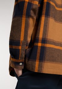 Camicia a quadri nei toni del marrone, arancione e blu navale. Presenta un polsino con bottone e una tasca laterale. Realizzata in tessuto testurizzato con un aspetto morbido.