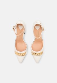 Escarpins blancs à bout pointu avec un accent chaîne dorée et une bride à la cheville. Finition en cuir lisse avec une découpe en forme de cœur sur la bride.