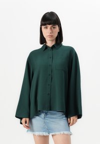 Mørkegrønn knapp-up skjorte med krage, lange ermer, en frontlomme og en avslappet passform, kombinert med en frynset denimskjørt.