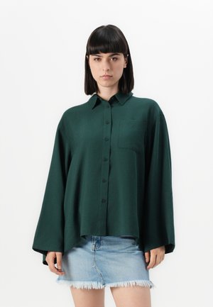Mørkegrønn knapp-up skjorte med krage, lange ermer, en frontlomme og en avslappet passform, kombinert med en frynset denimskjørt.