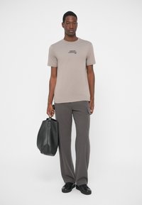 Man wearing beige Armani Exchange T-shirt, dark gray pants, black sneakers, holding large black leather bag against white background.