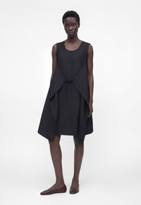 Μοντέλο με κοντά μαλλιά φοράει μαύρο αμάνικο φόρεμα με ασύμμετρο στρωματοποιημένο μπροστινό μέρος και μπορντό μπαλαρίνες, στέκεται μπροστά σε λευκό φόντο.