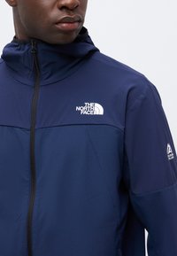 Άνδρας που φοράει μια σκούρα μπλε ζακέτα The North Face με φερμουάρ μπροστά και κουκούλα σε μονόχρωμο φόντο.
